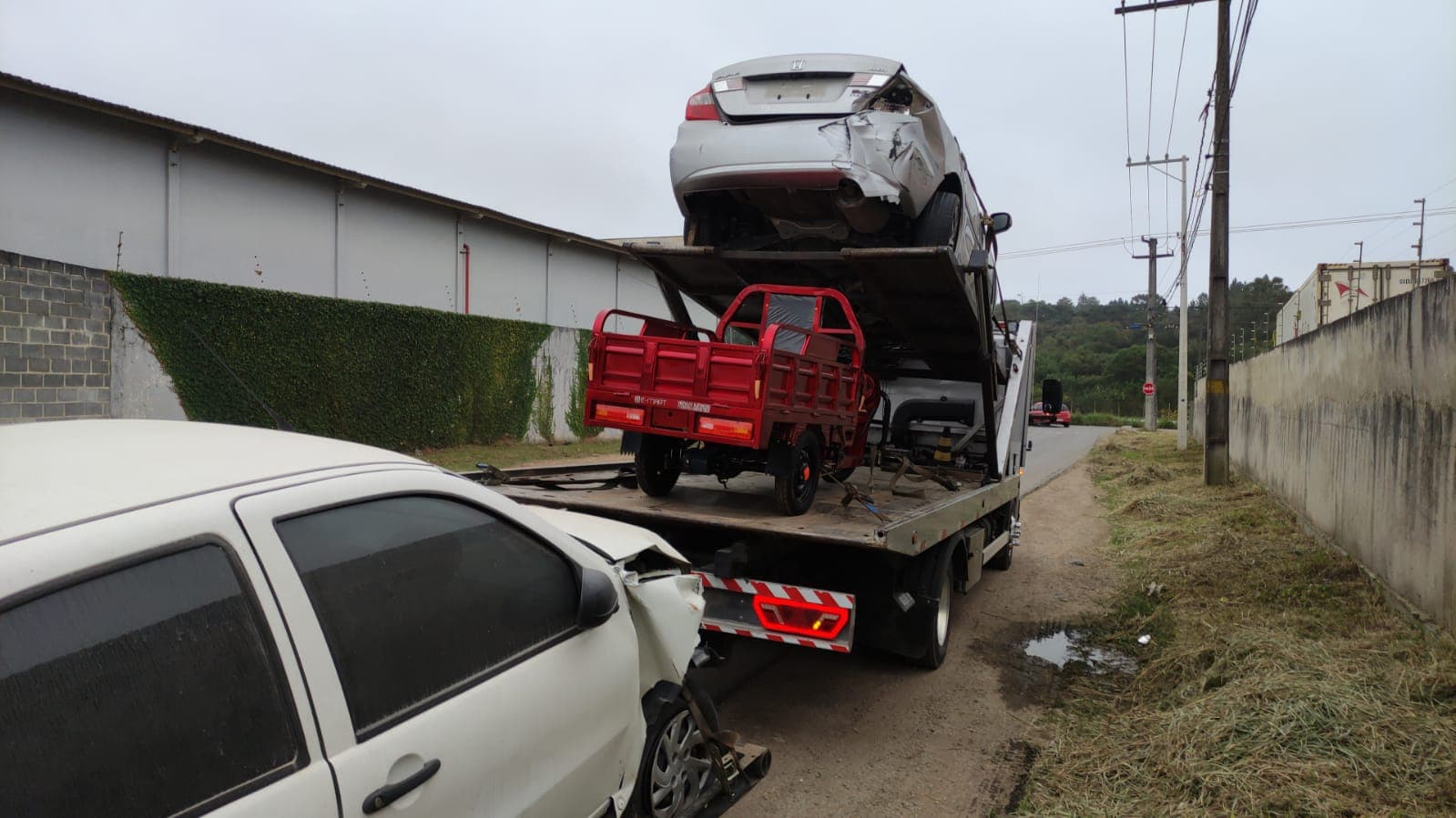 Uma foto mostrando a traseira do caminhão carregado com um carro em cima,um triciclo em baixo e um outro carro sendo arrastado